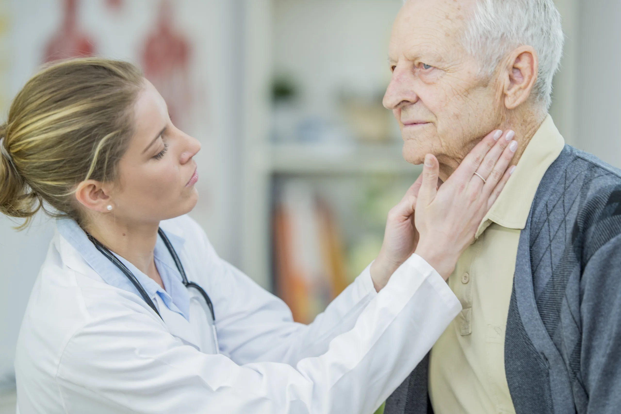 A Caucasian female doctor and elderly Caucasian man are indoors in a hospital. She is wearing doctor’s clothes and he is wearing casual clothing. She is using her hands to check his temples.