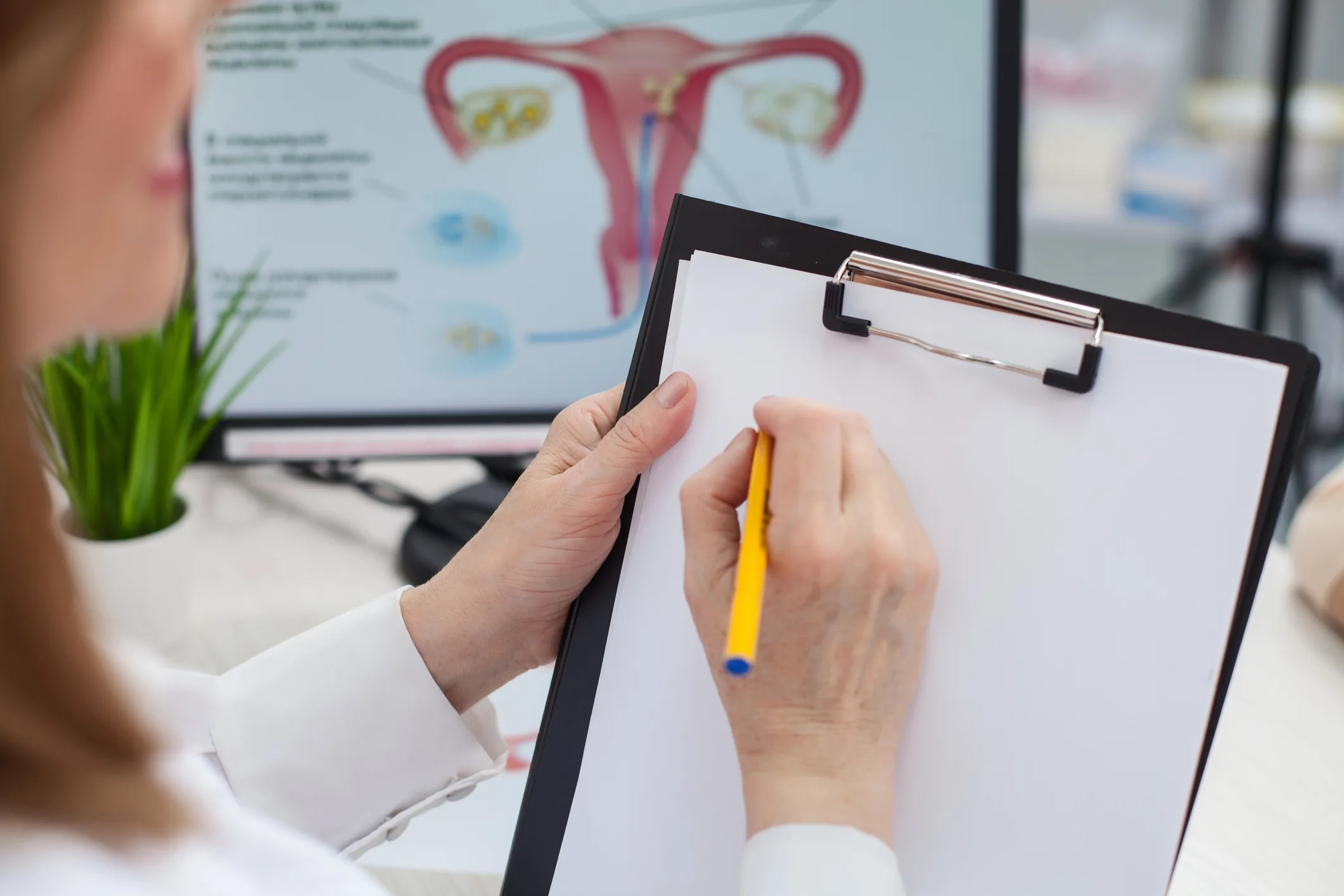 Close up of cheerful female gynecologist is writing a prescription. She is sitting at the desk and holding a folder of papers. The woman is smiling