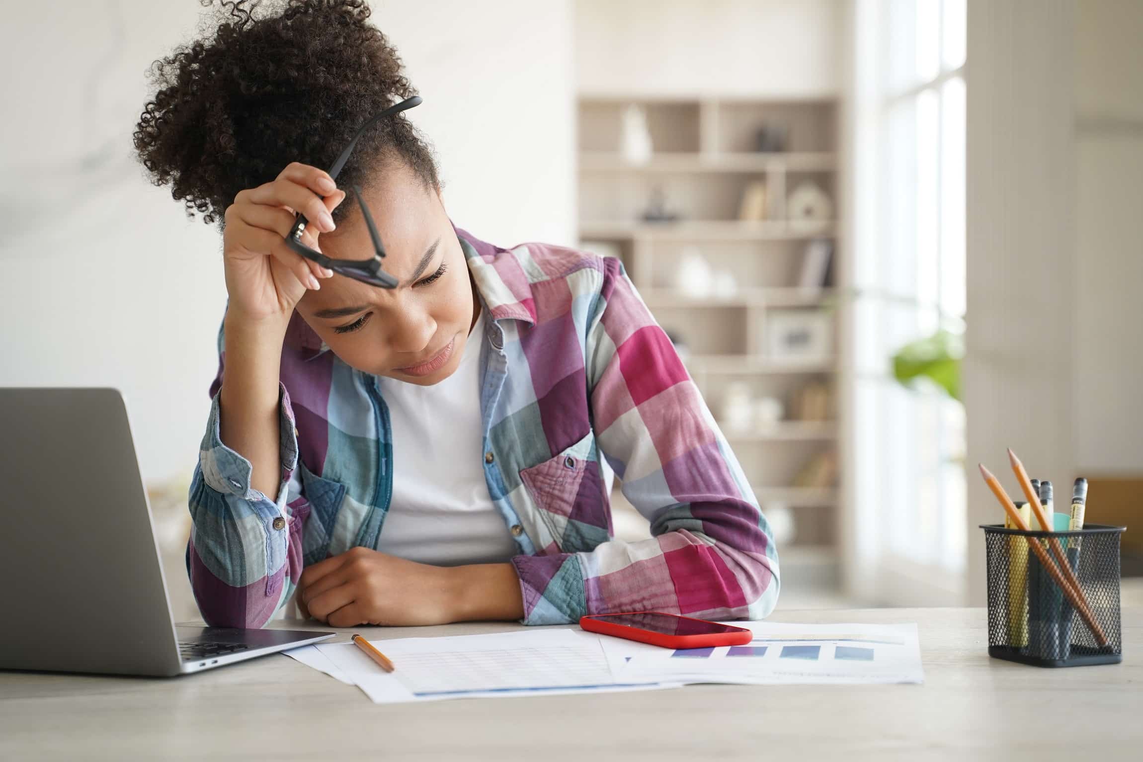 Stressed mixed race high school student girl studying with laptop at desk, preparing for exam. Puzzled teen schoolgirl student solves difficult task, doing homework learning at home. Academic stress.