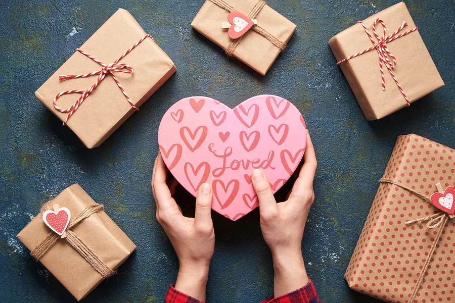 Close up on female hands holding a gift in a pink heart presents for valentine day, birthday, mother’s day. Flat lay. Symbol of love. Valentines day background with a gift boxes on concrete board.