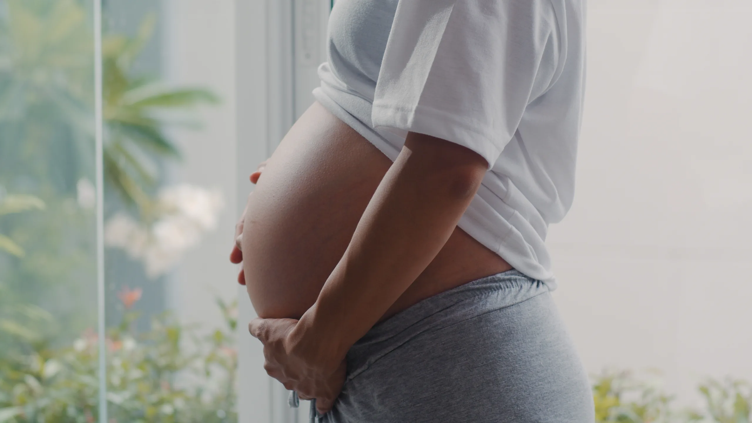 Young Asian Pregnant woman holding her belly talking with her child. Mom feeling happy smiling positive and peaceful while take care baby, pregnancy near window in living room at home concept.