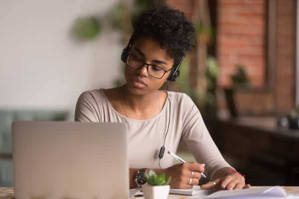 Focused mixed race woman wearing headphones watching webinar write notes study online with skype teacher, african female student learning language computer course on laptop listen translate lecture (Focused mixed race woman wearing headphones watching