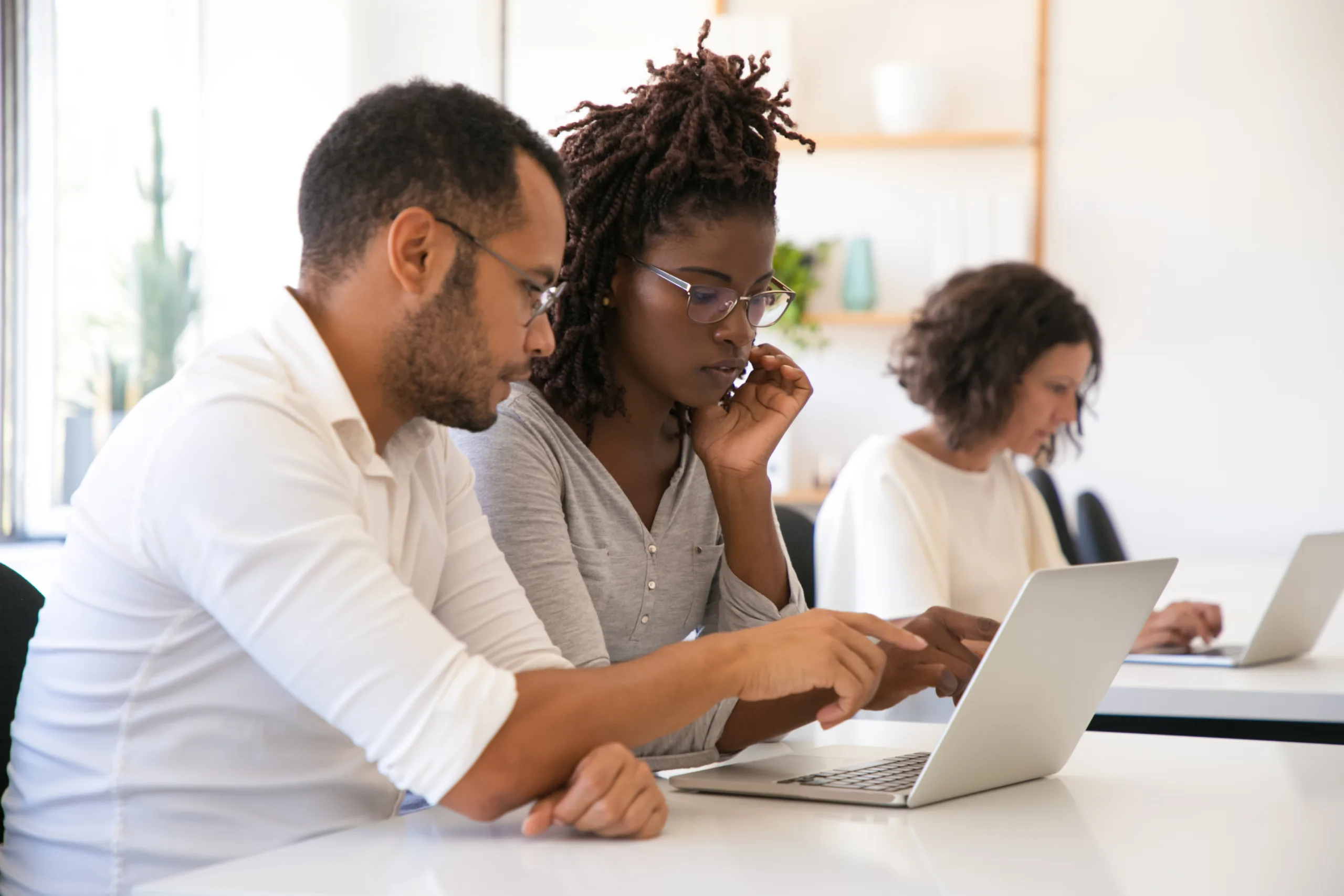 Instructor explaining corporate software specific to intern. Man and woman in casual sitting at desk in classroom, working on laptop, pointing at screen, talking. Education concept