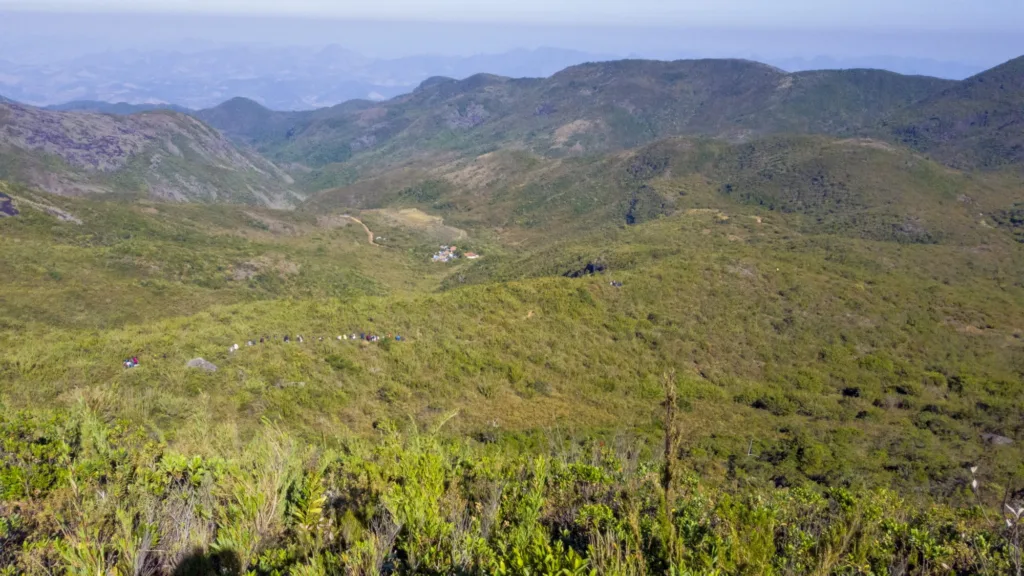 Trilha para o Pico da Bandeira com acampamento ao fundo e turistas descendo. Na imagem é possivel ver a vegetação típica do local.