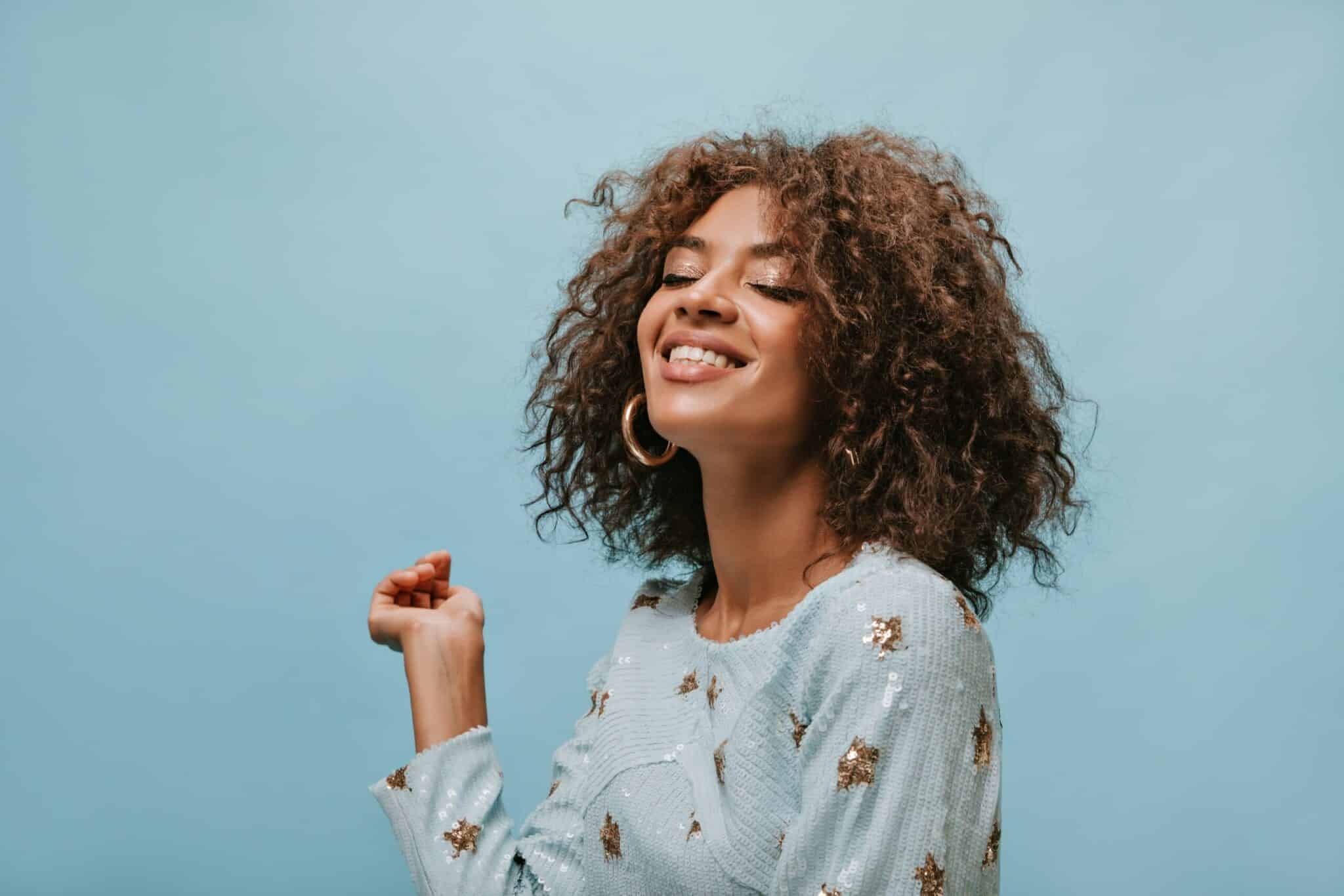 Charming woman with brunette short hair in stylish gold earrings and printed blue clothes smiling with closed eyes on isolated backdrop..