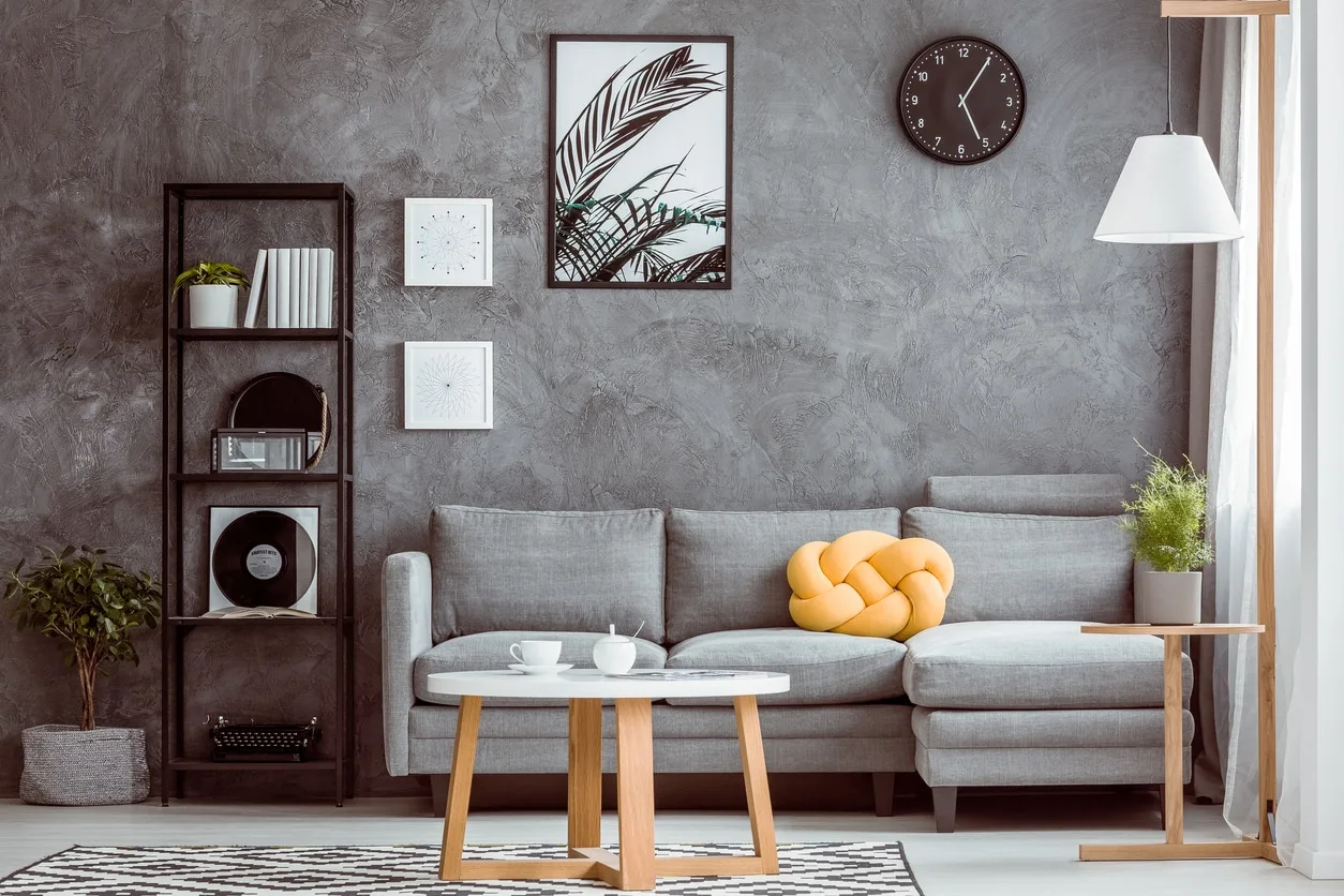 Black clock and posters on dark wall above grey settee with yellow pillow in cozy living room