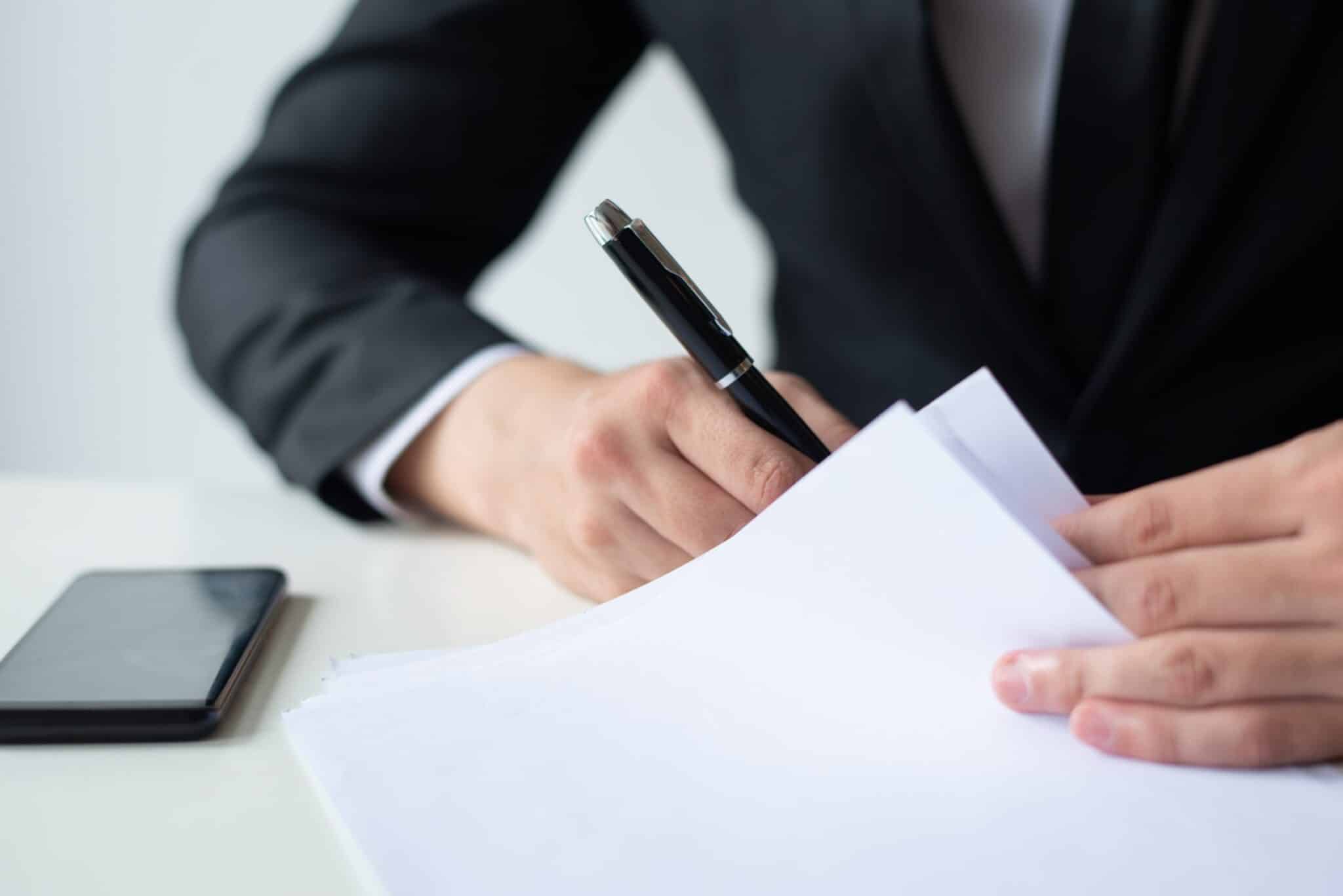 Closeup of business man signing document at office desk. Entrepreneur wearing suit and working. Contract concept. Cropped front view.