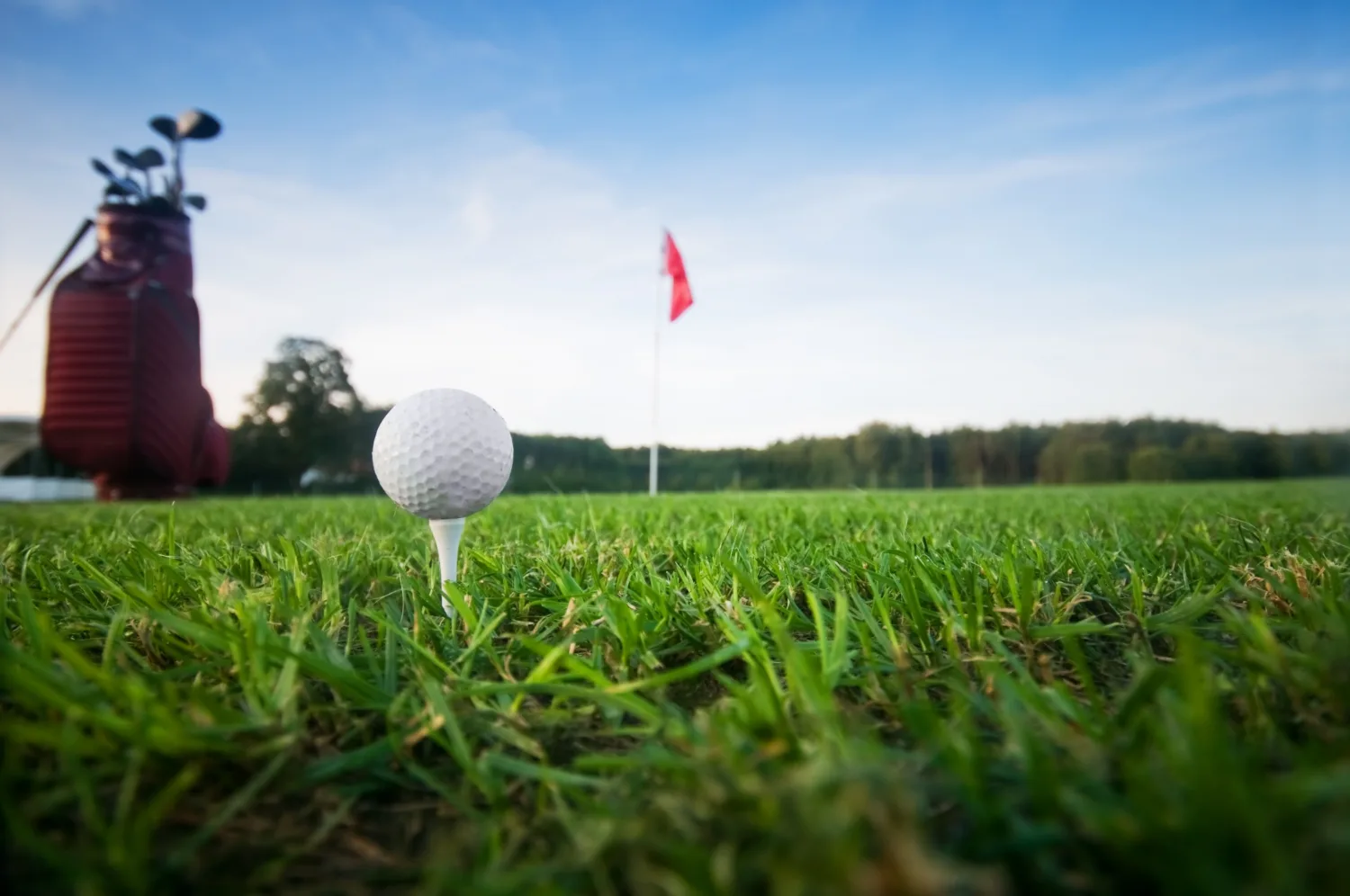 bola de golfe em destaque, sobre o suporte fincado na grama, à esquerda uma bolsa com tacos de golfe e o céu ao fundo