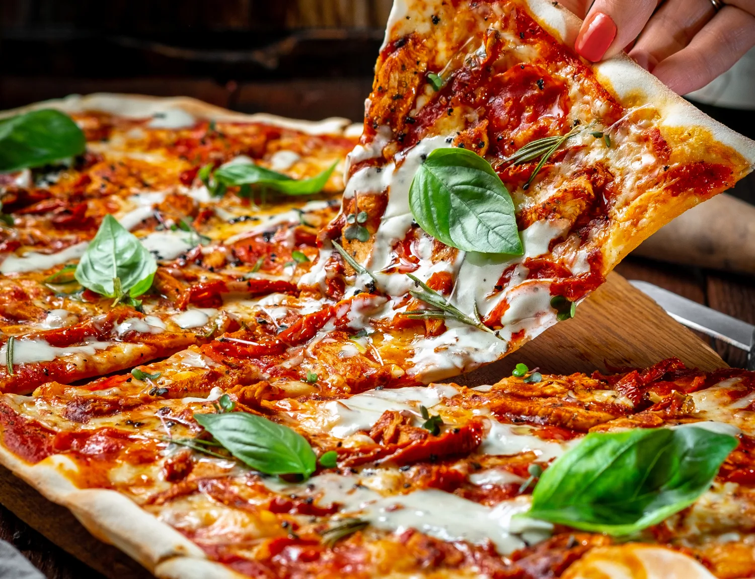 Hand takes a piece of Long pizza Margarita with sun-dried tomatoes and Basil on a wooden Board. Italian cuisine rustic style