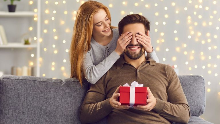 Mulher surpreendendo marido com presente, colocando as mãos no rosto dele.