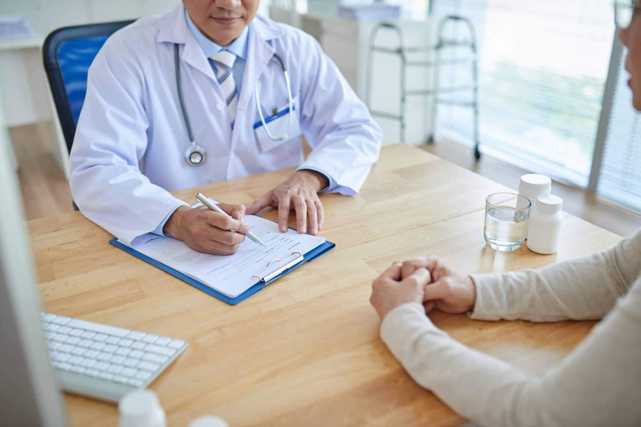 Close-up shot of male doctor filling in medical record, his patient sitting opposite him