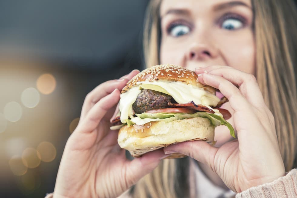 A young looks down, wide eyed,  about to eat a huge cheeseburger crammed with fillings.