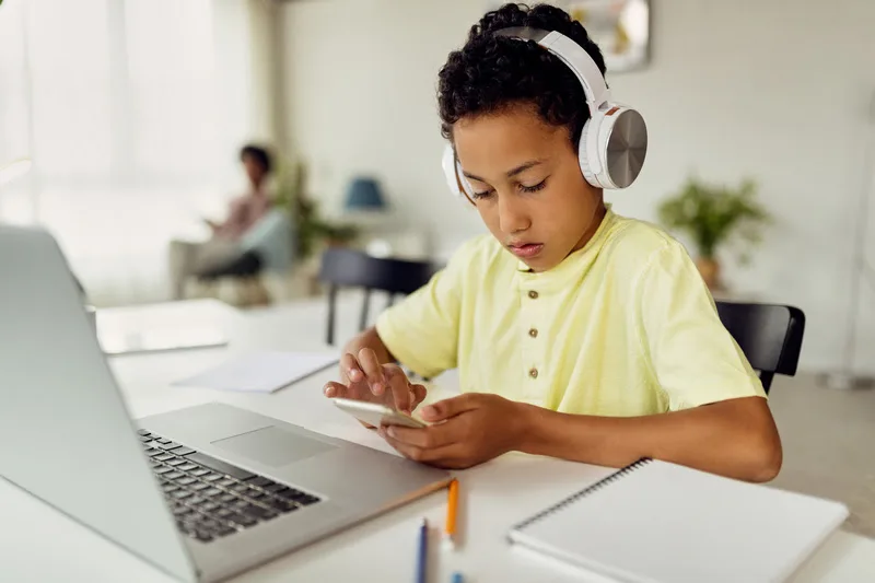 Small black boy wearing headphones and texting on mobile phone while learning at home.