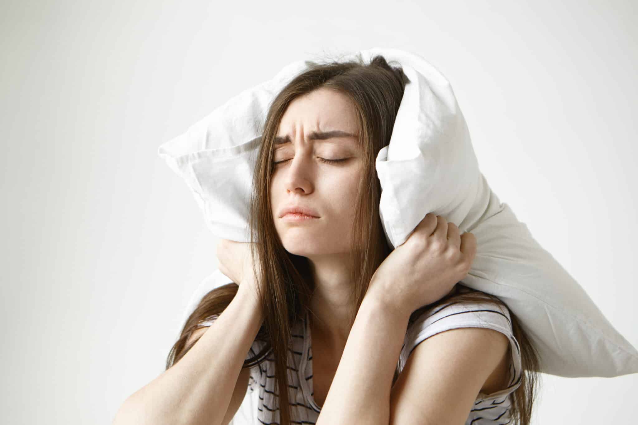 Pretty student girl in striped t-shirt trying to have some sleep, sitting in bedroom with pillow on her head, closing eyes, having tired painful look, irritated with annoying sound. Negative emotions