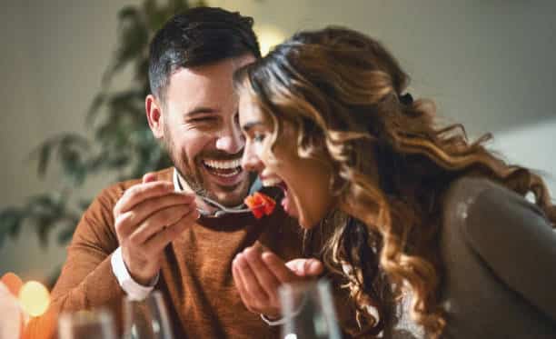 Closeup of mid 20's couple having fun during dinner party. The guy is feeding his girls with some chopped fruit, both laughing.