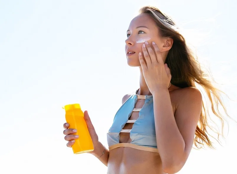 Charming woman in stylish swimsuit putting tanning cream on her face, sunbathing on the beach. Healthcare.