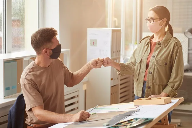 Warm toned portrait of two colleagues wearing masks bumping fists as contactless greeting in post pandemic office