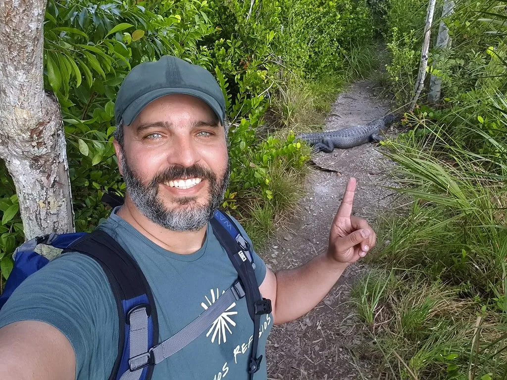 Observador da natureza tirando selfie silenciosa com um jacaré em uma trilha, demonstrando a importância da observação cuidadosa."Finalidade da Imagem: A imagem ilustra a prática de observação silenciosa na natureza, enfatizando a necessidade de não perturbar os animais selvagens durante a fotografia.