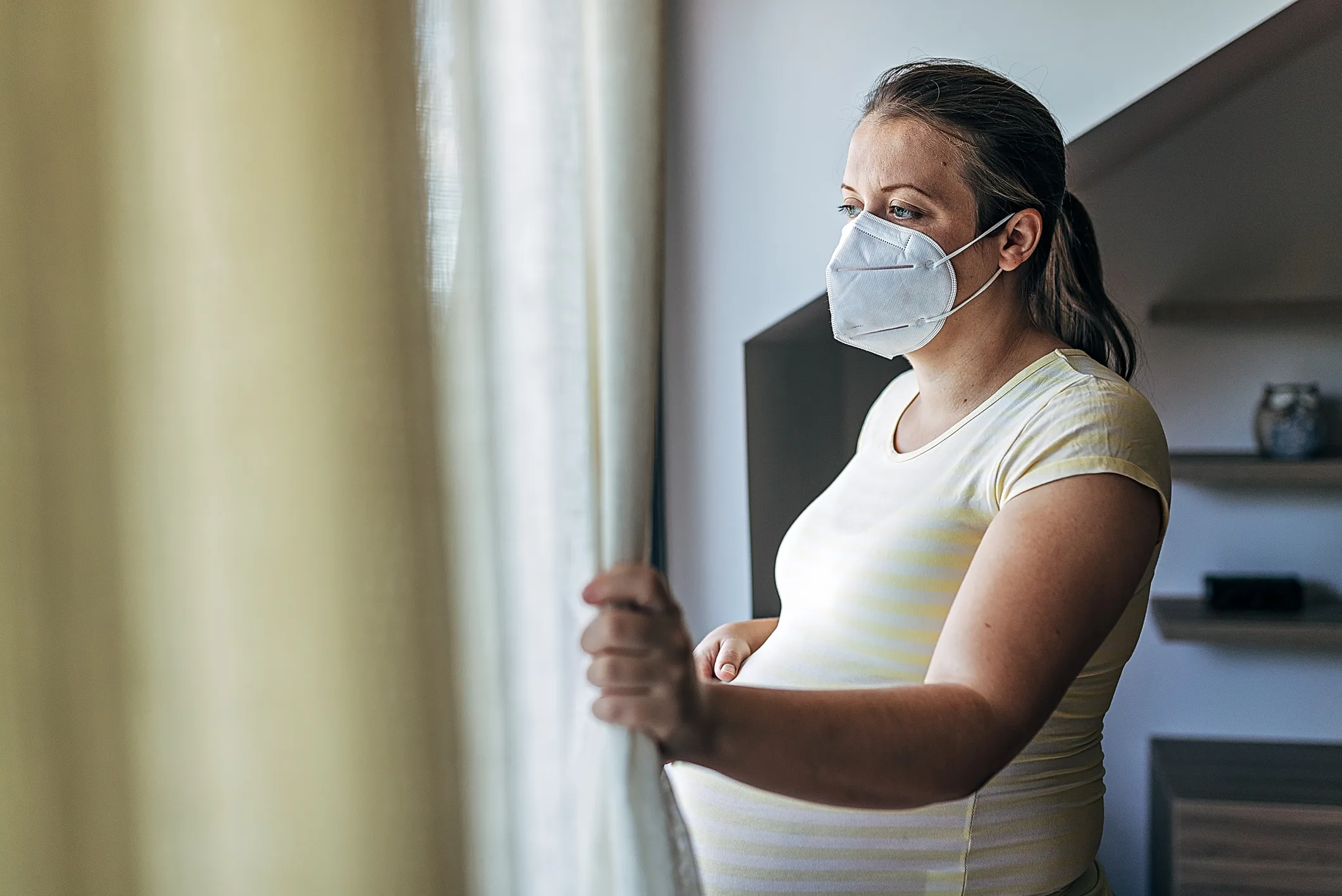 Pregnant woman with the face mask standing near window