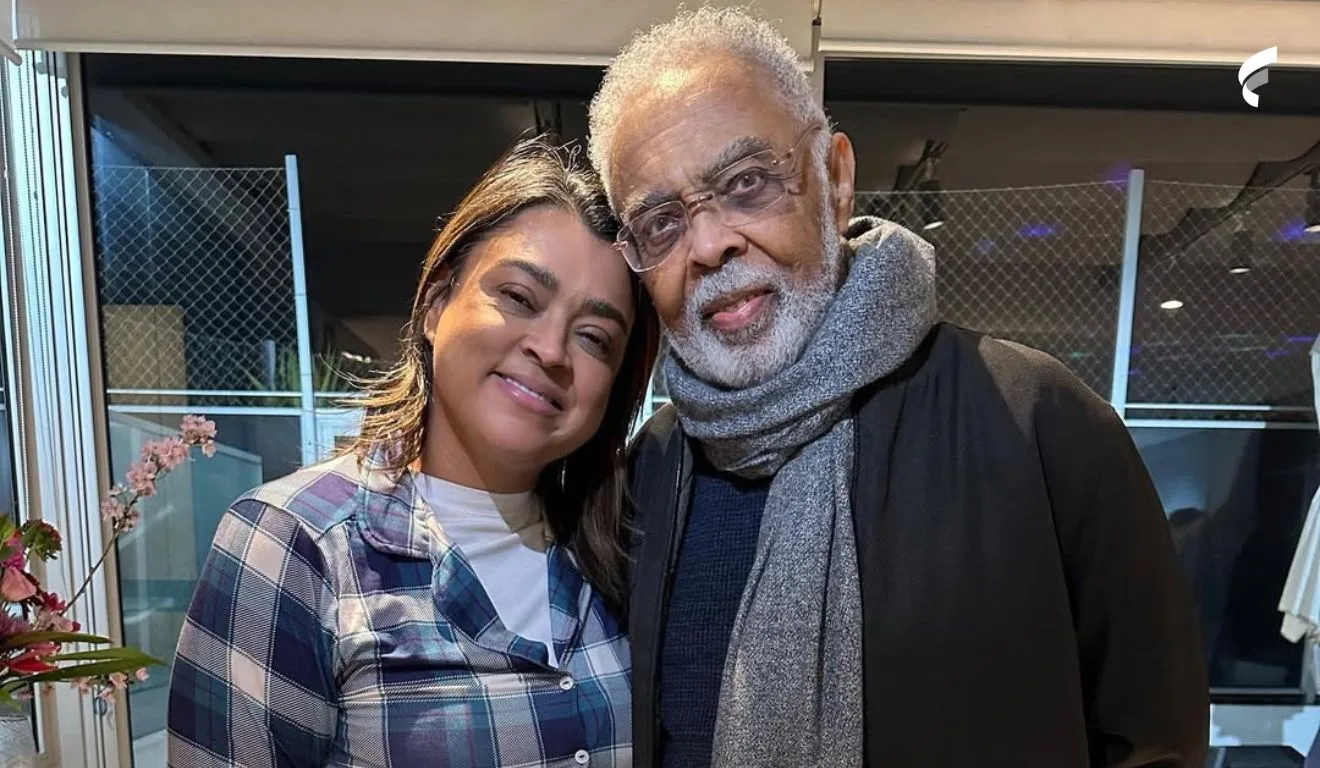 Preta Gil e Gilberto Gil aparecem abraçados e sorrindo para a foto. Ela usa uma blusa de manga longa xadrez e ele um casaco preto e um cachecol cinza