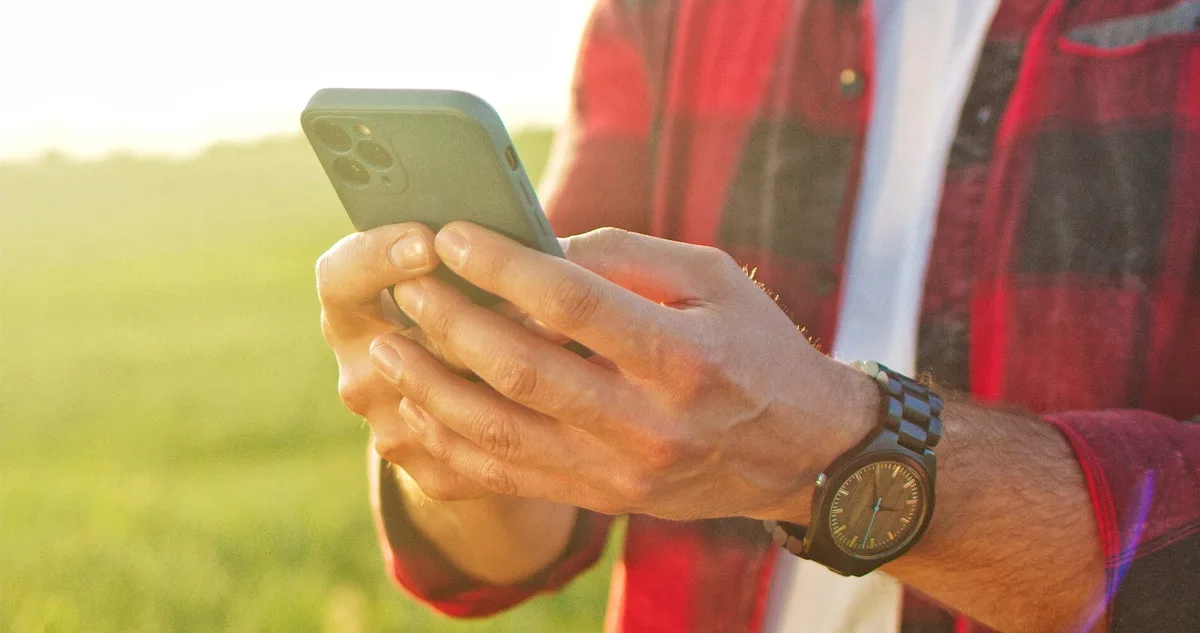 Cropped view of the optimistic caucasian farmer scientist in plaid shirt using smartphone mobile technology application while working at the field and smiling. Agriculture concept