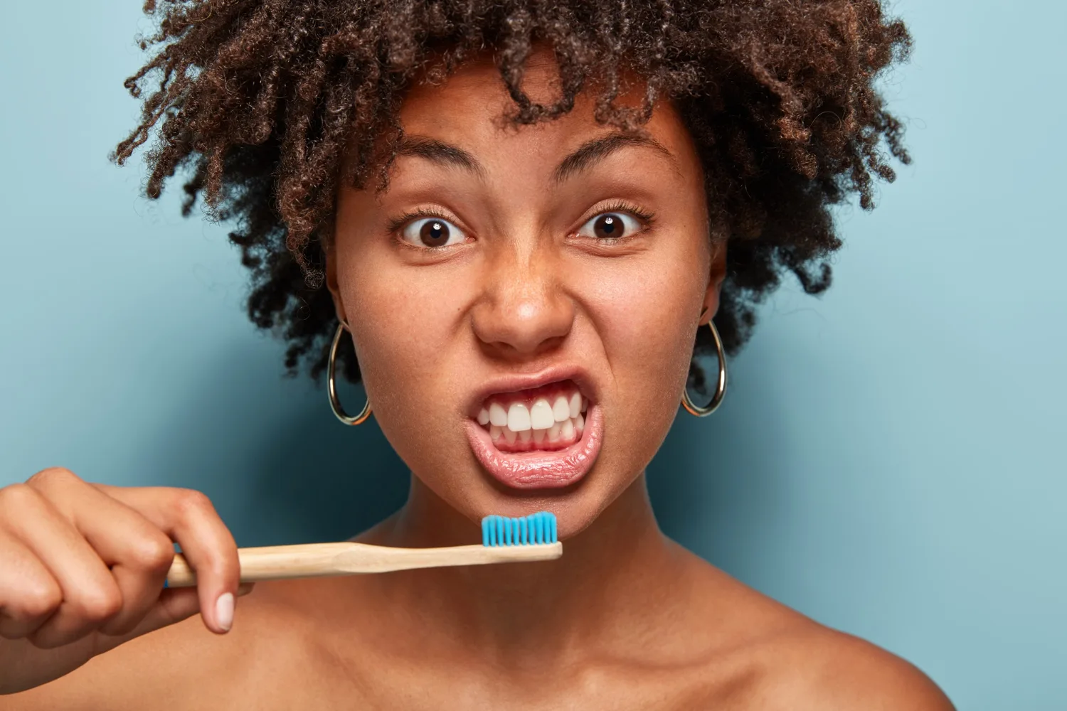 Jovem negra, de cabelos crespos castanhos mostra os dentes com uma escova de dentes com pasta em frente à boca, na posição de iniciar a escovação