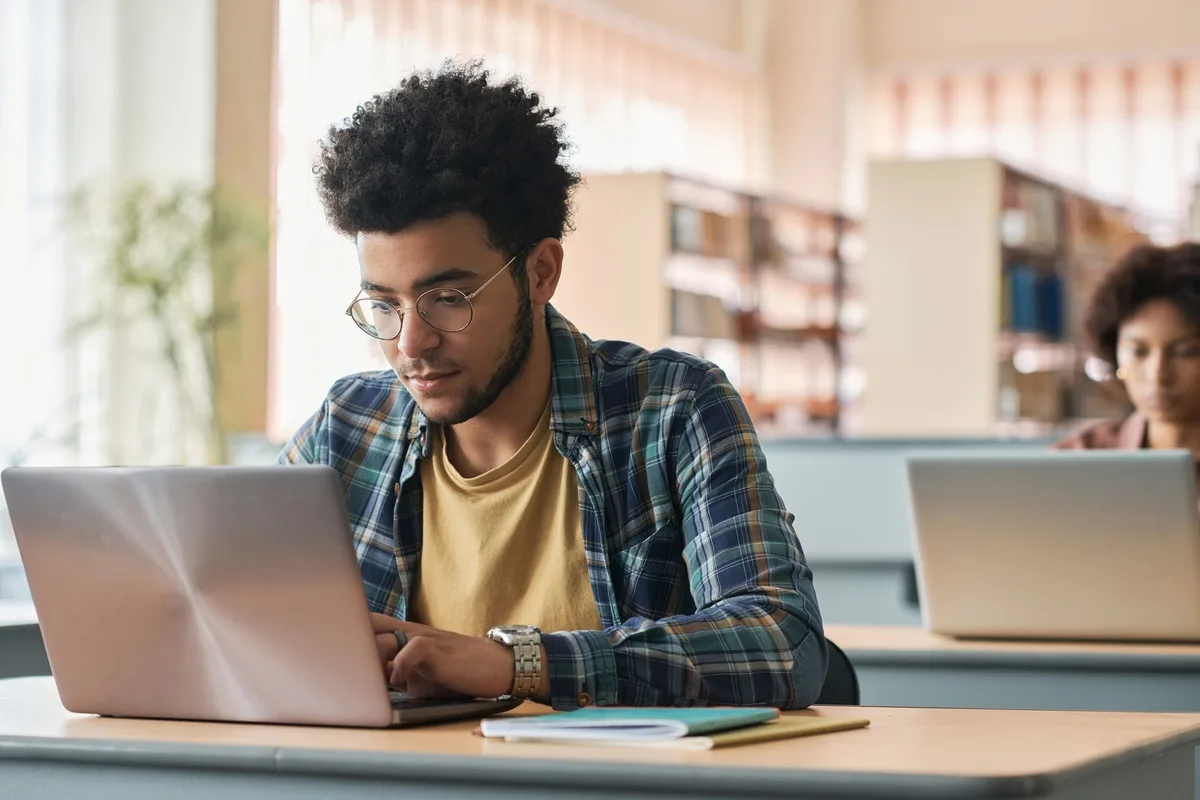 Young migrant sitting at desk and studying online with laptop with other students in class