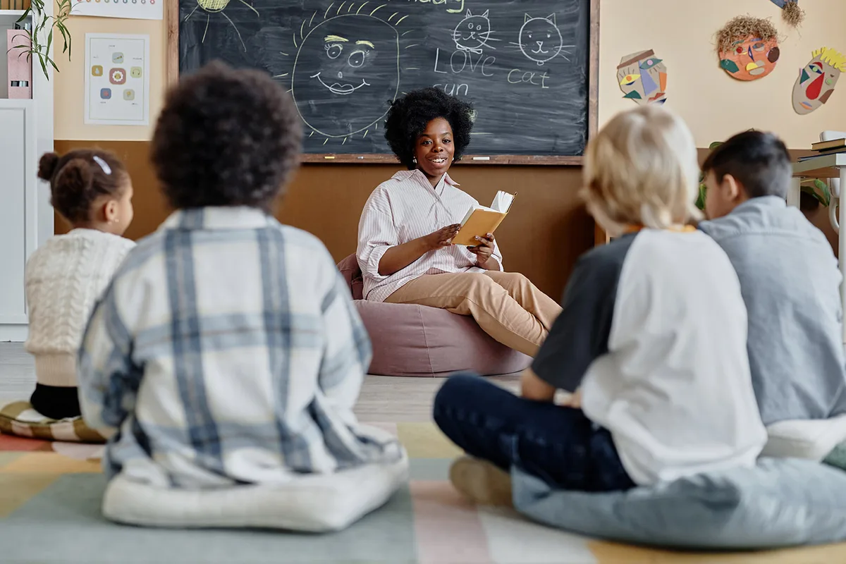 Young female teacher of Black ethnicity in bean bag chair reading book together with children sharing thoughts about story while sitting on floor in classroom