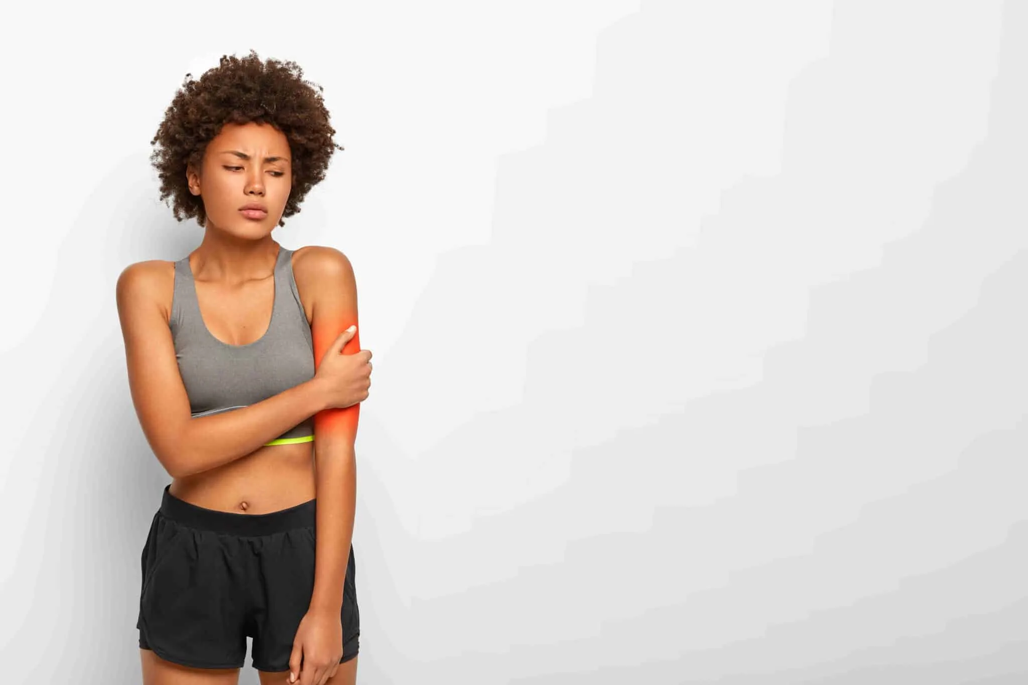 Horizontal view of upset Afro American woman touches arm, suffers from painful feelings, hurted hand during fitness training, dressed in casual top and shorts, poses indoor over white studio wall