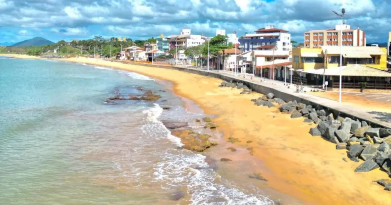 Uma cidade costeira com prédios e hotéis em Guarapari ao longo de uma estrada pavimentada próxima a uma praia de areia, com ondas chegando à costa sob um céu parcialmente nublado. Uma cidade costeira com prédios e hotéis em Guarapari ao longo de uma estrada pavimentada próxima a uma praia de areia, com ondas chegando à costa sob um céu parcialmente nublado.
