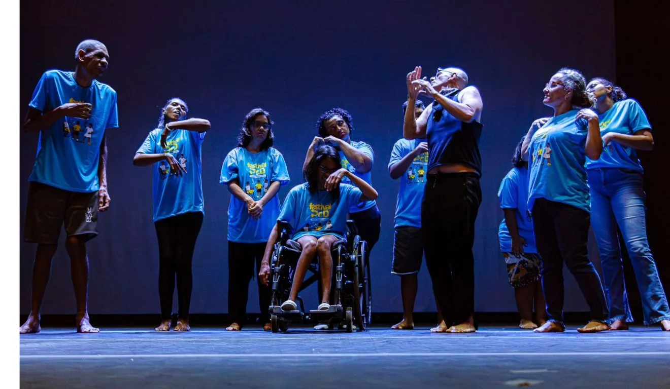 Dez artistas no palco usando uma camisa azul e calça jeans. No centro, uma delas está em uma cadeira de rodas. O resto está em pé e parecem dançar